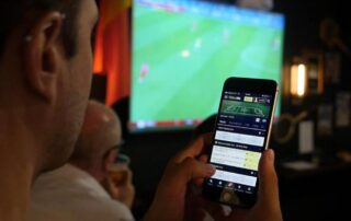 Person placing a live sports bet on a smartphone while watching a soccer match on a big screen.