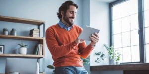 Smiling man in an orange sweater using a tablet in a home office.