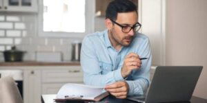 Man in glasses working on a laptop while reviewing documents at home.