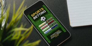 Smartphone displaying a sports betting app on a dark table, with a notepad and glass nearby.