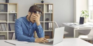 Frustrated man holding head in hand while using laptop at desk.