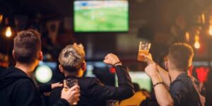 Group of men cheering with drinks while watching a live football match on a big screen in a bar