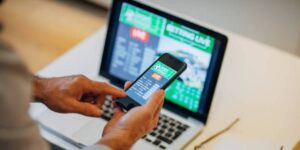 Hands using a sports betting app on a smartphone with live odds on a laptop in the background.