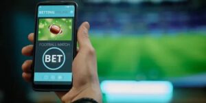 Close-up of a hand holding a smartphone with a football betting app open, ready to place a bet, with a stadium blurred in the background. 