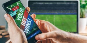A person holding a smartphone showing a live sports betting app displaying “Your Bet Wins!” with a football match streaming on a laptop in the background.