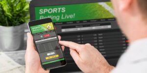 A person using a smartphone to place a live sports bet, with the same betting platform displayed on a laptop screen in the background.