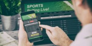 Man placing a live sports bet on a mobile phone while viewing betting odds on a laptop screen.