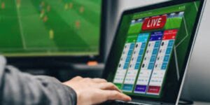 Person using a laptop with live sports betting software while watching a match on a screen.