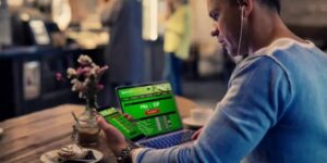 Man in a café using both his phone and laptop to view online sports betting odds.