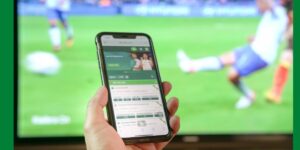 Hand holding a smartphone showing football betting odds in front of a live match on television