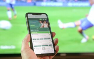 Hand holding a smartphone showing football betting odds in front of a live match on television
